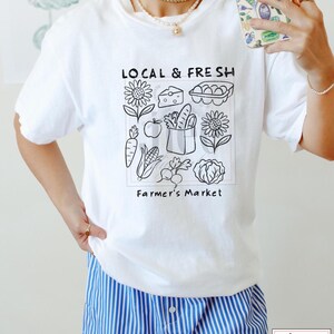 May include: White t-shirt with a black graphic that reads "LOCAL & FRESH" above a square illustration of farmer's market produce. The illustration includes sunflowers, cheese, eggs, carrots, apples, bread, corn, radishes, and a cabbage. The text "Farmer's Market" is below the illustration.