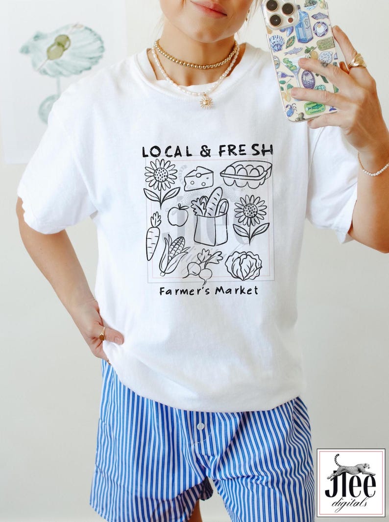 May include: White t-shirt with a black graphic that reads "LOCAL & FRESH" above a square illustration of farmer's market produce. The illustration includes sunflowers, cheese, eggs, carrots, apples, bread, corn, radishes, and a cabbage. The text "Farmer's Market" is below the illustration.