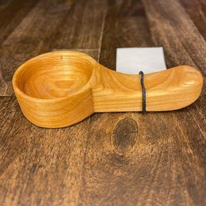 May include: A handcrafted wooden scoop with a rounded bowl and a handle. The scoop is made of light-colored wood, possibly maple or birch, and has a smooth, polished finish. A black string is attached to the handle. The scoop is designed for kitchen use.