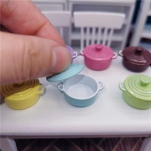 May include: A collection of miniature round Dutch oven pots in various colors including yellow, light blue, pink, brown, and light green. Each small pot features two side handles and a matching removable lid, displayed on a white surface.