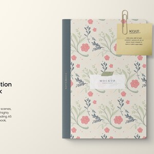 Puede incluir: Una maqueta de cuaderno de composición con un diseño floral en rosa y verde. El cuaderno está cerrado y tiene un lomo azul. Un clip de papel dorado sujeta una nota adhesiva amarilla con la palabra "MOCKUP" escrita en ella. También se incluye un bolígrafo y un lápiz en la imagen.