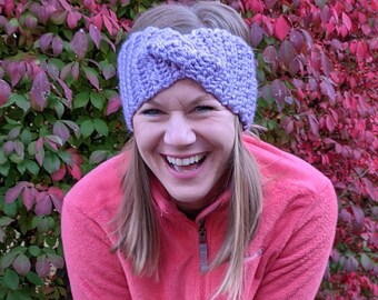 May include: A woman wearing a purple knitted headband with a twist design. She is smiling and wearing a pink fleece jacket.