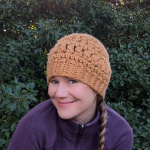 Crochet beanie hat with textured stitches in a yellow-mustard color being worn by a light skinned woman in a purple jacket.