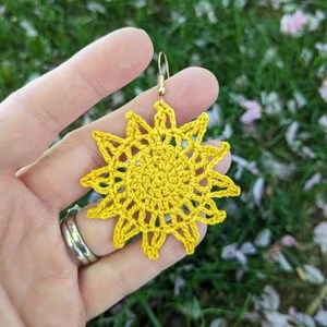 May include: A handmade, yellow crochet sun earring with a gold-colored hook. The sunburst design features a detailed, openwork pattern. The earring is held against a blurred background of green grass and pink flower petals.