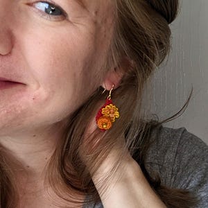 Puede incluir: Un par de pendientes colgantes con flores de ganchillo en tonos rojos, naranjas y amarillos.