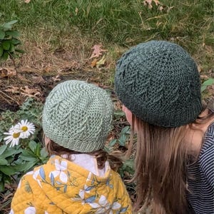 Könnte beinhalten: Zwei Personen tragen grüne Strickmützen mit einem Blatt-Muster. Die Person im Vordergrund trägt eine gelbe Jacke mit einem Blumenmuster. Die Person im Hintergrund trägt ein gestreiftes Hemd.