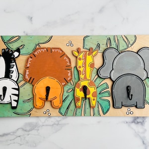 May include: Wooden wall hook featuring a zebra, lion, giraffe, and elephant. Each animal has a hook for hanging items. The background has a jungle theme with green leaves and white dots. The animals are painted in their natural colors.