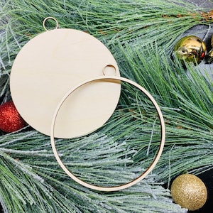 Peut inclure: Deux ornements en bois non finis, l'un rond et l'autre un cercle, avec une boucle de ficelle pour accrocher. Les ornements sont sur un lit de branches de pin vertes avec des ornements dorés et rouges.