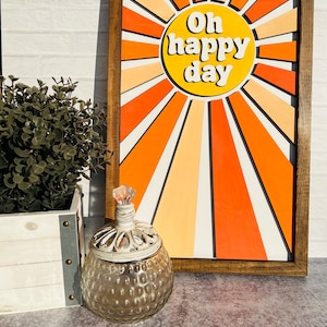 May include: A wooden framed wall art piece with a yellow sun and orange and white rays. The sun has the text "Oh happy day" in white.