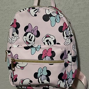 K&ouml;nnte beinhalten: Rosa Rucksack mit einem sich wiederholenden Muster von Minnie Mouse Gesichtern mit Schleifen in verschiedenen Farben. Der Rucksack hat eine Rei&szlig;verschlusstasche vorne, einen oberen Griff und goldfarbene Rei&szlig;verschl&uuml;sse. Die Tr&auml;ger sind rosa.