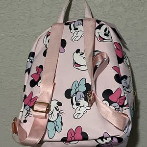K&ouml;nnte beinhalten: Rosa Rucksack mit einem sich wiederholenden Muster von Minnie Mouse in verschiedenen Posen mit bunten Schleifen. Der Rucksack hat rosa Tr&auml;ger und einen oberen Griff, mit goldfarbener Hardware. F&uuml;r den Transport von Essentials konzipiert.