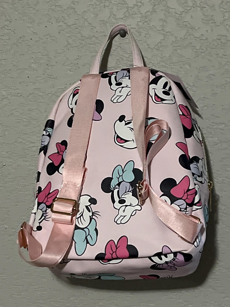 K&ouml;nnte beinhalten: Rosa Rucksack mit einem sich wiederholenden Muster von Minnie Mouse in verschiedenen Posen mit bunten Schleifen. Der Rucksack hat rosa Tr&auml;ger und einen oberen Griff, mit goldfarbener Hardware. F&uuml;r den Transport von Essentials konzipiert.