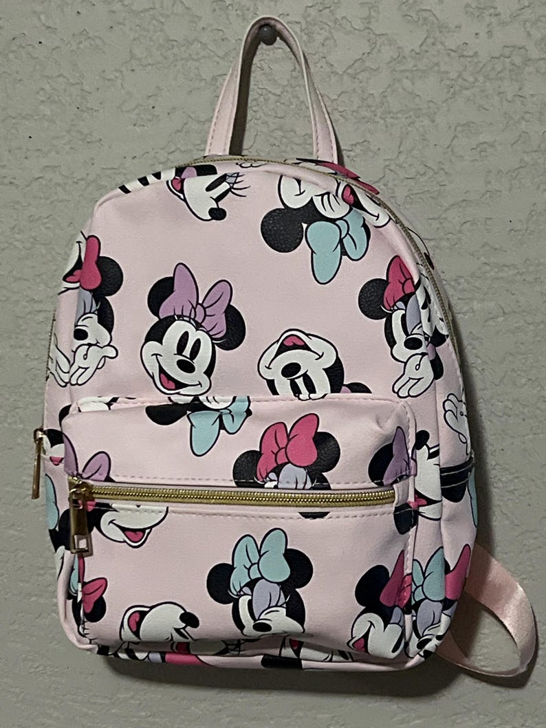 K&ouml;nnte beinhalten: Rosa Rucksack mit einem sich wiederholenden Muster von Minnie Mouse Gesichtern mit Schleifen in verschiedenen Farben. Der Rucksack hat eine Rei&szlig;verschlusstasche vorne, einen oberen Griff und goldfarbene Rei&szlig;verschl&uuml;sse. Die Tr&auml;ger sind rosa.
