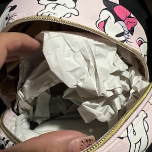 K&ouml;nnte beinhalten: Kleiner, runder rosa Rucksack mit einem sich wiederholenden Minnie Mouse-Muster. Er hat einen goldenen Rei&szlig;verschluss und einen oberen Griff. Der Innenraum ist mit wei&szlig;em Papier gef&uuml;llt. Die Tasche ist klein.