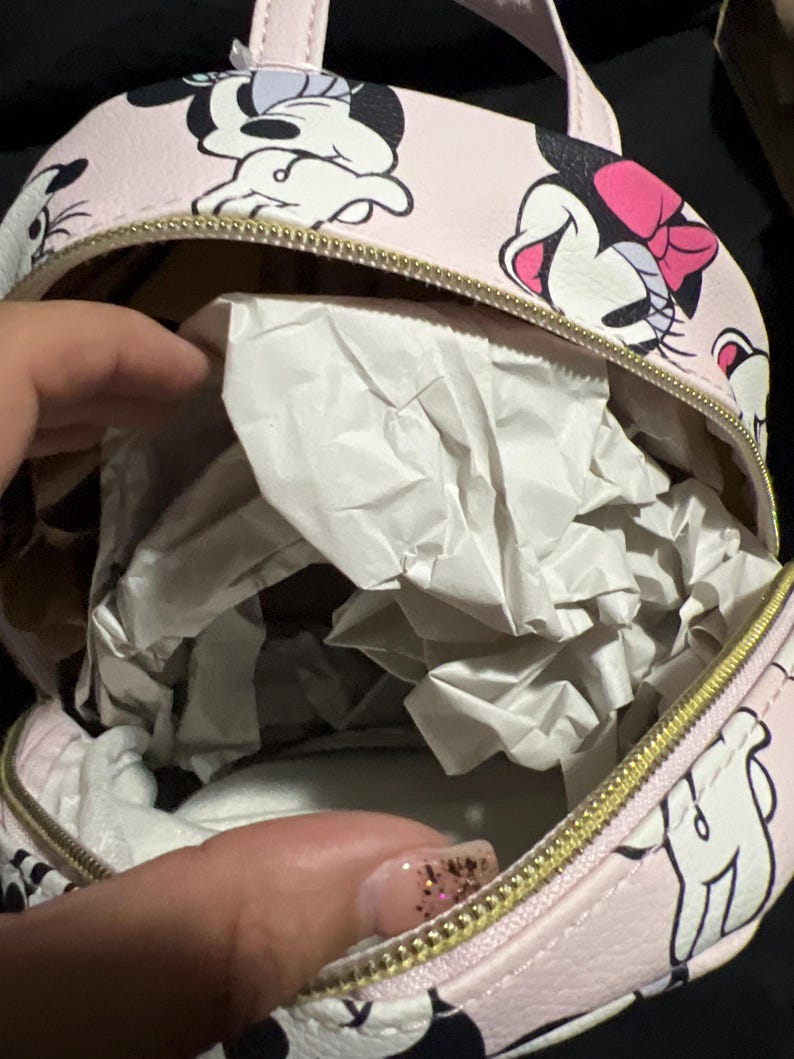 K&ouml;nnte beinhalten: Kleiner, runder rosa Rucksack mit einem sich wiederholenden Minnie Mouse-Muster. Er hat einen goldenen Rei&szlig;verschluss und einen oberen Griff. Der Innenraum ist mit wei&szlig;em Papier gef&uuml;llt. Die Tasche ist klein.