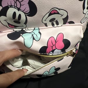 K&ouml;nnte beinhalten: Rosa Rucksack mit einem sich wiederholenden Muster von Minnie Mouse. Der Rucksack hat eine Rei&szlig;verschluss-Vordertasche und einen goldenen Rei&szlig;verschluss. Minnie Mouse wird mit schwarzem Kopf, rosa Schleife und blauer Schleife dargestellt.