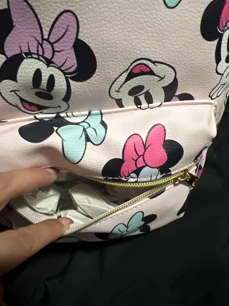K&ouml;nnte beinhalten: Rosa Rucksack mit einem sich wiederholenden Muster von Minnie Mouse. Der Rucksack hat eine Rei&szlig;verschluss-Vordertasche und einen goldenen Rei&szlig;verschluss. Minnie Mouse wird mit schwarzem Kopf, rosa Schleife und blauer Schleife dargestellt.