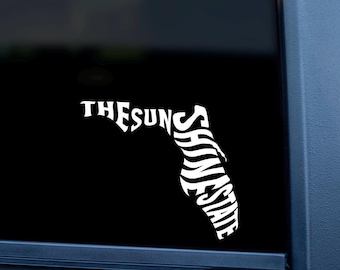May include: White vinyl decal on a car window. The decal is in the shape of the state of Florida, with the words "THE SUNSHINE STATE" in a stylized font. The decal is on a black background.