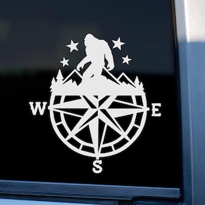 A white vinyl decal of a compass with a silhouette of Bigfoot standing on a mountain range inside the compass. The compass has the letters N, S, E, and W around the outside of the compass. The decal is on a black car window.