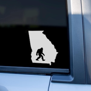 May include: A black and white vinyl decal featuring the outline of the state of Georgia with a silhouette of Bigfoot inside. The decal is affixed to the window of a vehicle.