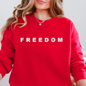 May include: A red sweatshirt with the word "FREEDOM" printed in white block letters. The sweatshirt is a solid red color and has long sleeves. The person in the image is wearing blue jeans.