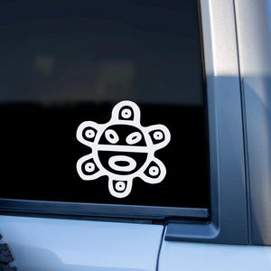 May include: White vinyl decal featuring a stylized sun design, applied to a vehicle window. The sun symbol has a face-like appearance with circular and oval shapes. The decal is a simple, clean design.