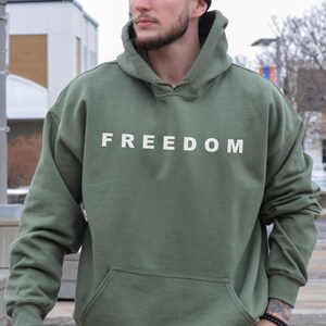 “Military green FREEDOM hoodie worn outdoors by man, minimalist white text sweatshirt.”