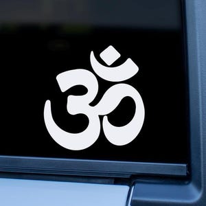 May include: White Om symbol sticker applied to a car window. The Om symbol is a sacred sound and spiritual icon in Hinduism, Buddhism, and Jainism. The sticker is a simple, clean design, with a glossy finish, and is applied to a black window.