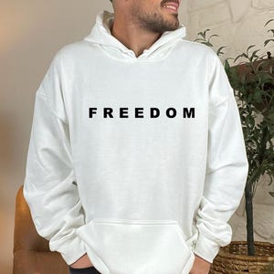 May include: White hoodie with the word "FREEDOM" printed in black across the chest. The hoodie has a front pocket and a hood. The person is wearing a green cap and black jeans.