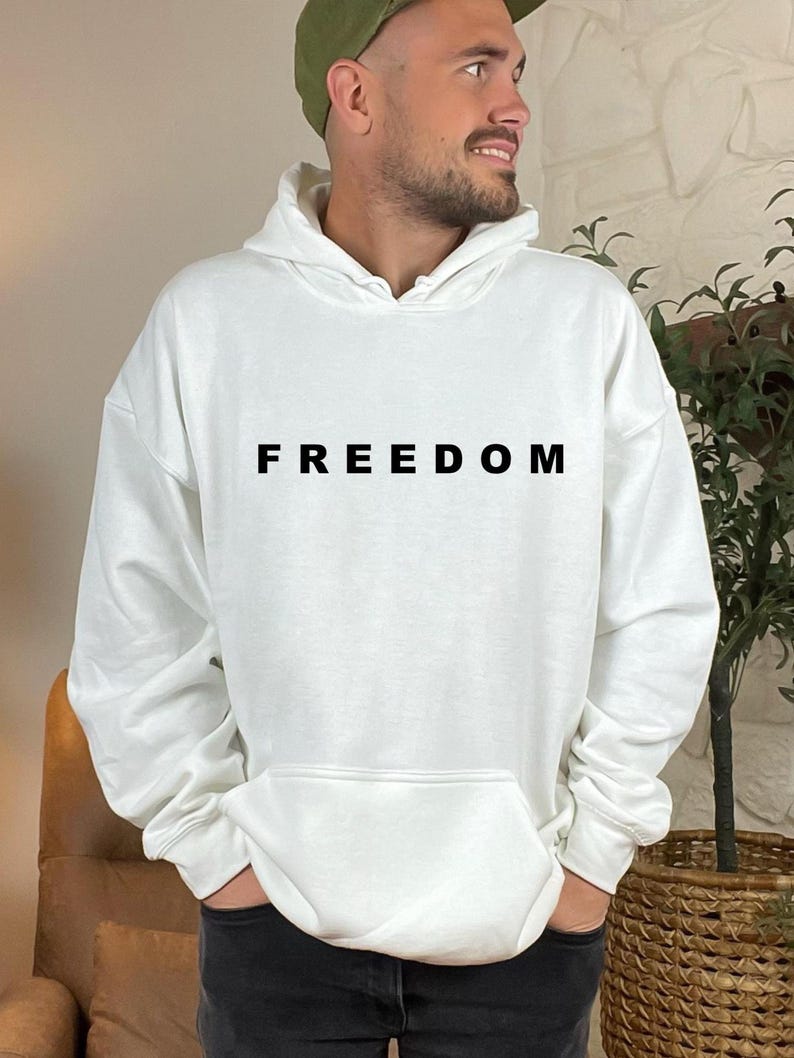 May include: White hoodie with the word "FREEDOM" printed in black across the chest. The hoodie has a front pocket and a hood. The person is wearing a green cap and black jeans.
