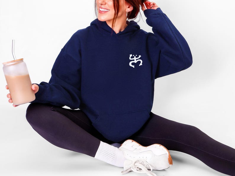May include: A navy blue hoodie with a white graphic on the chest. The hoodie is paired with black leggings and white sneakers. A glass tumbler with a straw and a light brown beverage is held in the hand.