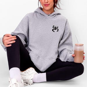 May include: A light gray hoodie with a black graphic on the chest. The hoodie is paired with black leggings and white sneakers. A clear glass with a brown beverage is held in the hand.
