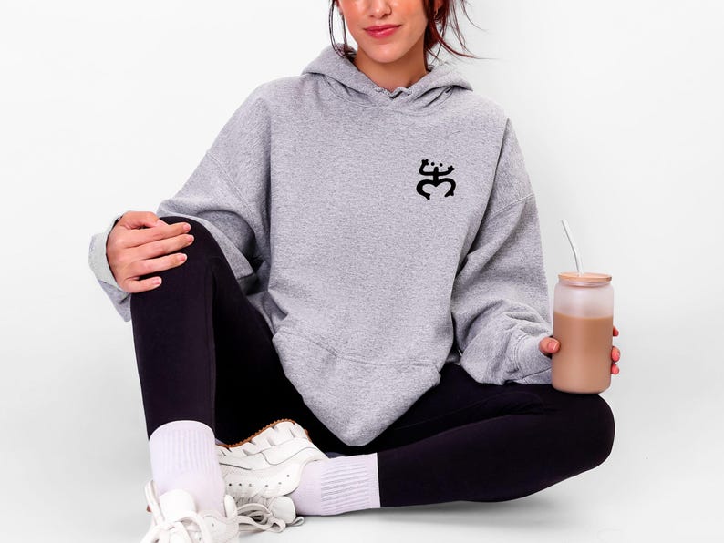 May include: A light gray hoodie with a black graphic on the chest. The hoodie is paired with black leggings and white sneakers. A clear glass with a brown beverage is held in the hand.