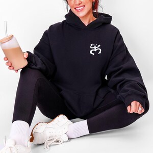 May include: Black hoodie with a white graphic on the chest. The person is wearing black leggings, white socks, and white sneakers. They are holding a glass tumbler with a wooden lid and a straw, filled with a light brown beverage.