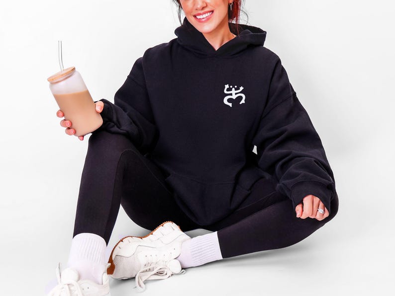 May include: Black hoodie with a white graphic on the chest. The person is wearing black leggings, white socks, and white sneakers. They are holding a glass tumbler with a wooden lid and a straw, filled with a light brown beverage.