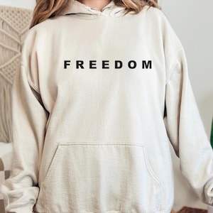 May include: A beige hoodie with the word "FREEDOM" printed in black across the chest. The hoodie has a kangaroo pocket and a drawstring hood. The fabric appears soft and comfortable, suitable for casual wear.