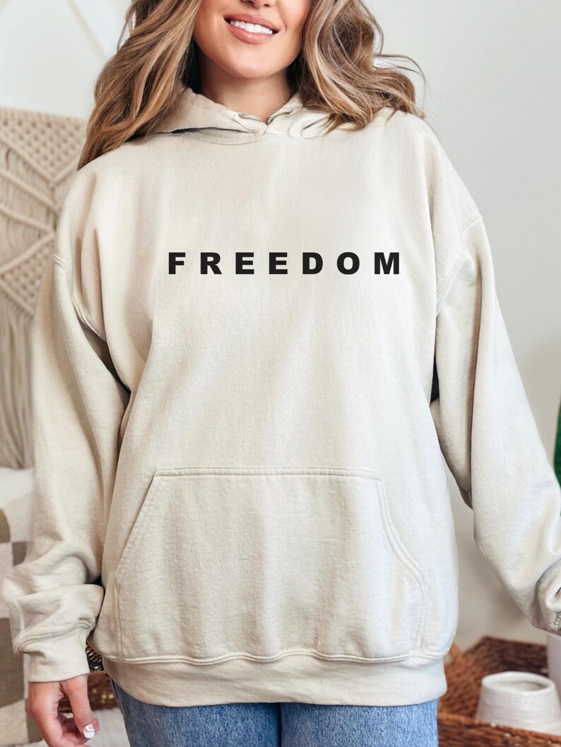 May include: A beige hoodie with the word "FREEDOM" printed in black across the chest. The hoodie has a kangaroo pocket and a drawstring hood. The fabric appears soft and comfortable, suitable for casual wear.