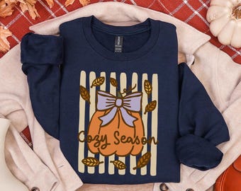 Puede incluir: Una sudadera azul marino de cuello redondo con un gráfico de una calabaza naranja con un lazo morado claro, rodeada de hojas marrones sobre un fondo de rayas verticales crema, con el texto Cozy Season escrito en letra marrón.