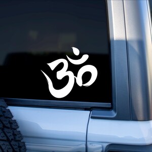 May include: White Om symbol decal on a black background, affixed to the rear window of a white vehicle. The Om symbol is a stylized, cursive design, representing a sacred sound and spiritual icon.