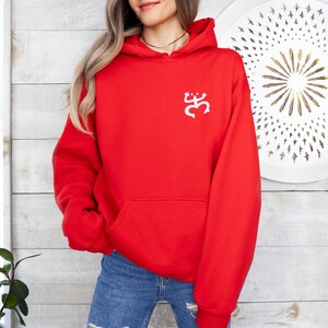 May include: A bright red hooded sweatshirt with a white graphic on the upper left chest. The sweatshirt has a kangaroo pocket and a drawstring hood. The person is wearing blue jeans. The background is a white and light brown wooden wall.