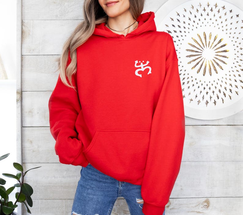 May include: A bright red hooded sweatshirt with a white graphic on the upper left chest. The sweatshirt has a kangaroo pocket and a drawstring hood. The person is wearing blue jeans. The background is a white and light brown wooden wall.