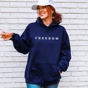 May include: Navy blue hoodie with the word "FREEDOM" in white block letters. The hoodie has a front pocket and a drawstring hood. The person in the image is wearing a white baseball cap and holding a clear glass.