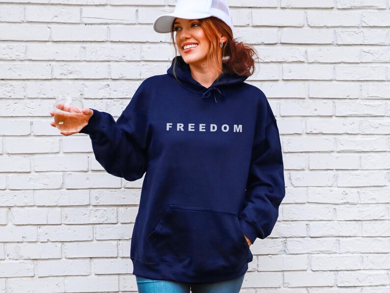 May include: Navy blue hoodie with the word "FREEDOM" in white block letters. The hoodie has a front pocket and a drawstring hood. The person in the image is wearing a white baseball cap and holding a clear glass.
