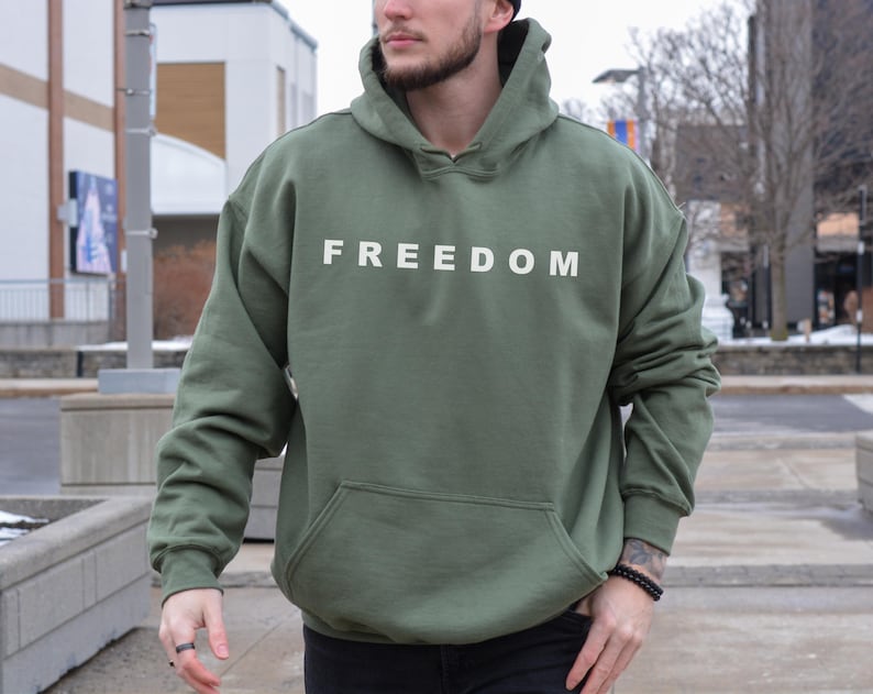 “Military green FREEDOM hoodie worn outdoors by man, minimalist white text sweatshirt.”