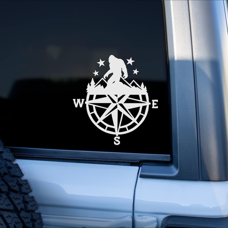 A white vinyl decal of a compass with a silhouette of Bigfoot standing on a mountain range inside the compass. The compass has the letters N, S, E, and W around the outside of the compass. The decal is on a black car window.