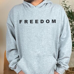 May include: A light gray hoodie with the word "FREEDOM" printed in black across the chest. The hoodie has a front pocket and a hood. The person is wearing black jeans.