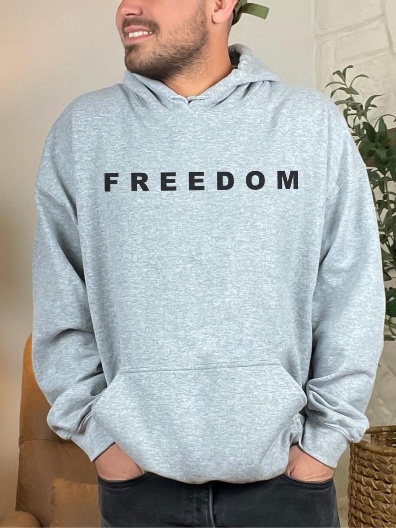 May include: A light gray hoodie with the word "FREEDOM" printed in black across the chest. The hoodie has a front pocket and a hood. The person is wearing black jeans.