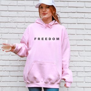 May include: A light pink hoodie with the word "FREEDOM" printed in black across the chest. The hoodie has a front pocket and a drawstring hood. The person in the image is wearing a white baseball cap and holding a clear glass.