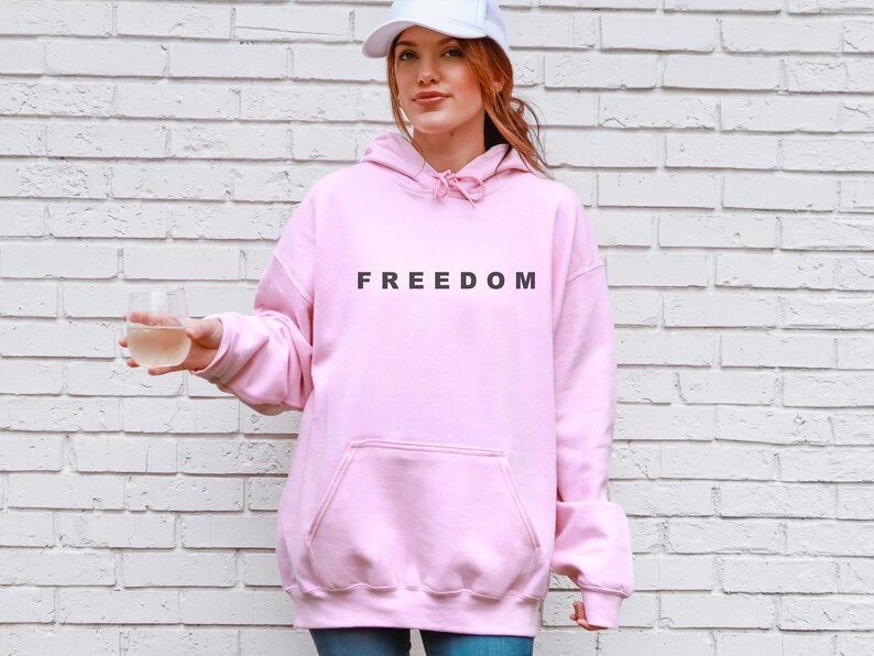 May include: A light pink hoodie with the word "FREEDOM" printed in black across the chest. The hoodie has a front pocket and a drawstring hood. The person in the image is wearing a white baseball cap and holding a clear glass.