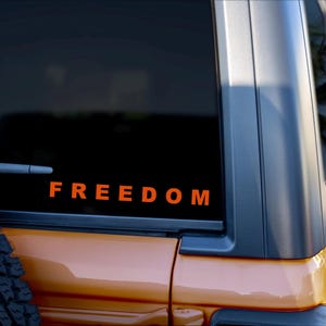 May include: Orange "FREEDOM" decal on the rear window of a vehicle. The vehicle is a light orange color with a black window frame. A spare tire is visible on the left side of the vehicle.